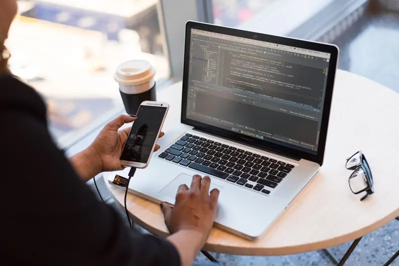 Persona programando en un computador portátil mientras revisa su teléfono